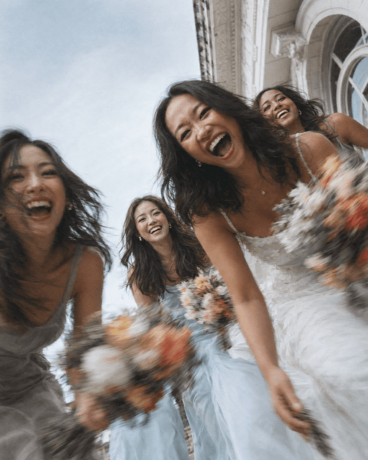 A bride and her friends rushing towards the camera laughing, bouquets swinging