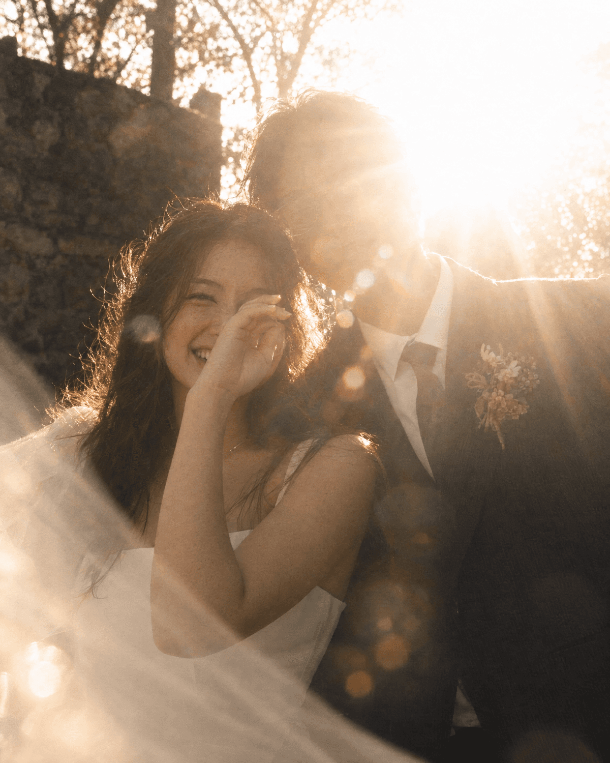 A bride laughing in low golden sunlight, her veil drifting across the frame