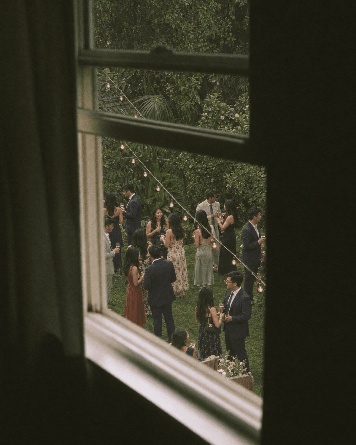 Guests seen through a dark window frame, gathered on the lawn under string lights